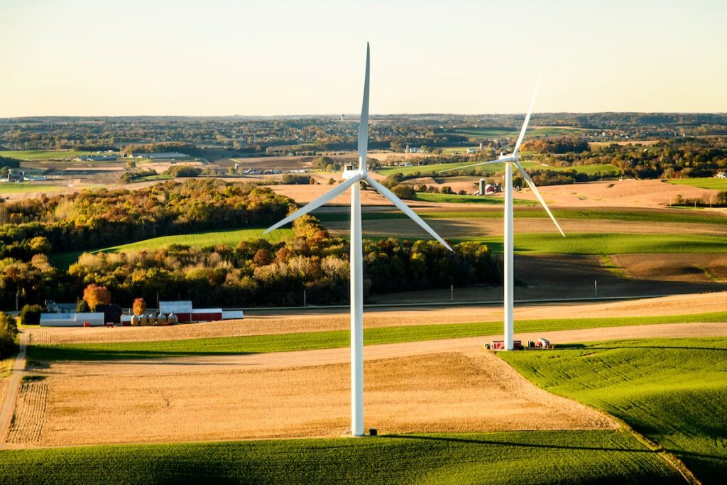 Two large wind turbines stand in an open rural landscape with fields, trees, and a few farm buildings under a clear sky.