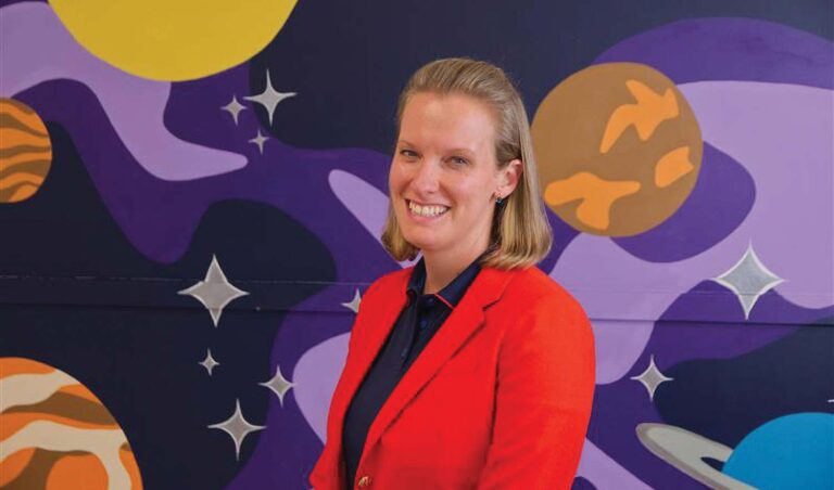 A bright classroom with large windows, small tables, and blue ceiling panels shaped like clouds.