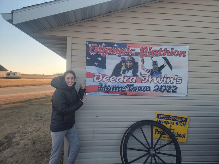 A biathlete wearing blue and red USA race gear skis on snow with a rifle on their back, against a background of trees.