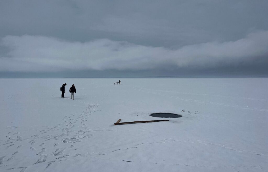 Several people stand on a vast snow-covered frozen lake near a circular hole in the ice, with footprints and wooden planks scattered nearby under a cloudy sky.
