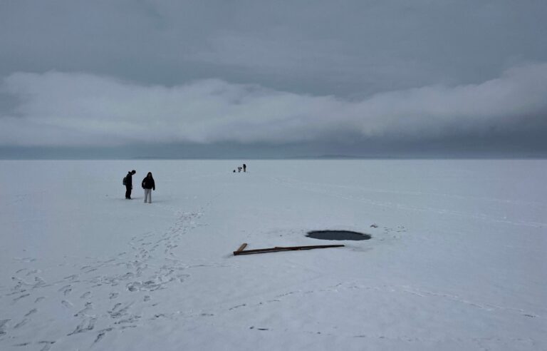 Several people stand on a vast snow-covered frozen lake near a circular hole in the ice, with footprints and wooden planks scattered nearby under a cloudy sky.