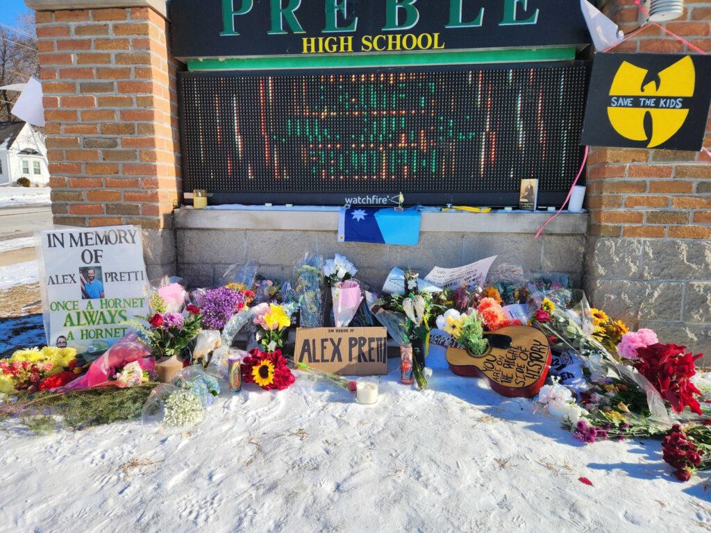 A memorial with flowers, candles, signs, and posters is arranged in front of a high school sign for Alex Pretti, with snow covering the ground.