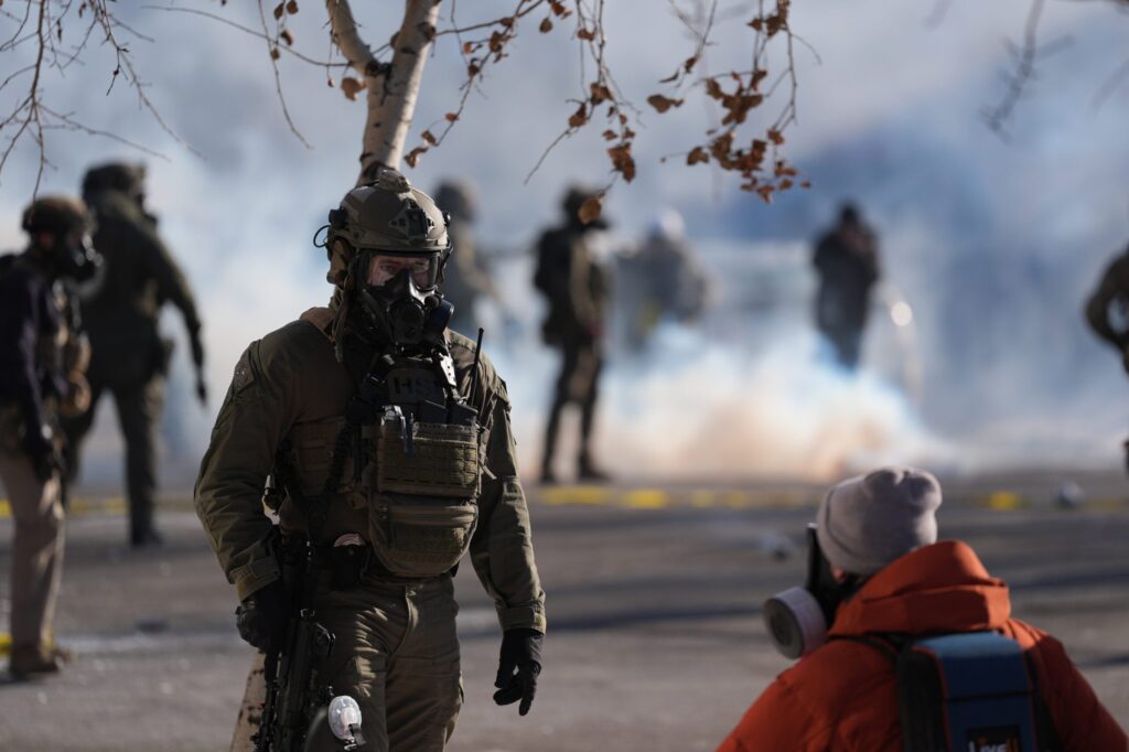 A group of law enforcement officers in tactical gear stand on a city street near stores, with police tape visible in the background.