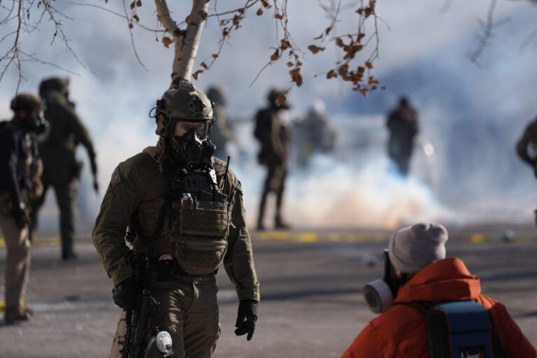 A group of law enforcement officers in tactical gear stand on a city street near stores, with police tape visible in the background.