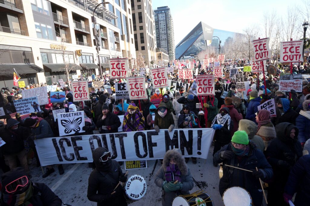 A large crowd gathers outdoors holding signs that say ICE OUT! and a banner reading ICE OUT OF MN! in front of urban buildings on a winter day.