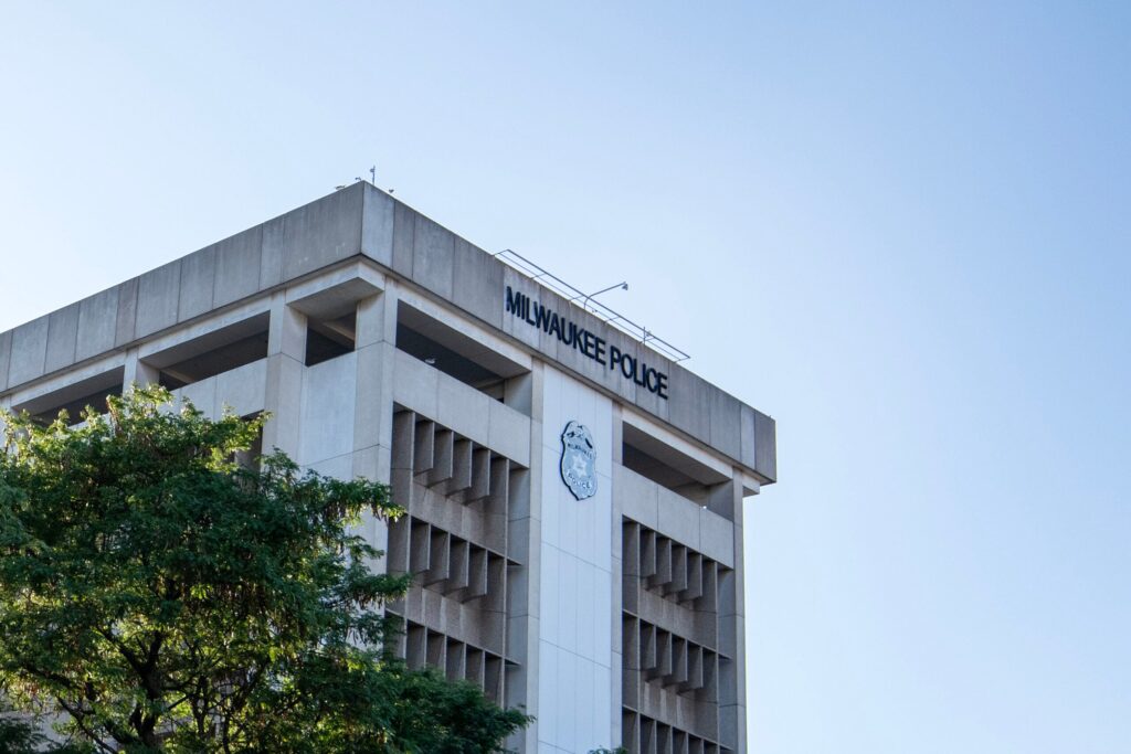 The image shows the exterior of the Milwaukee Police Department building with its name and emblem visible on the upper section. A tree partially obscures the lower part.
