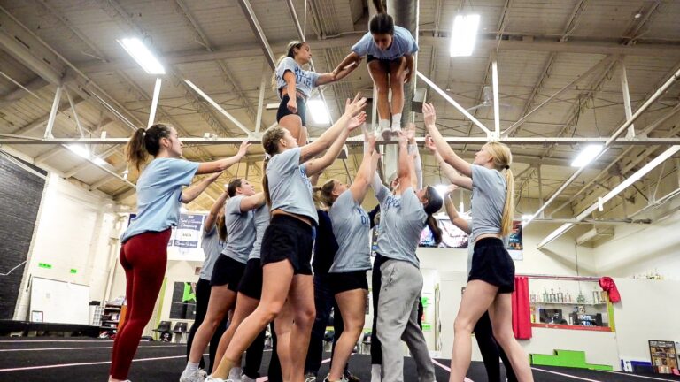 A group of cheerleaders in matching uniforms stand in a circle with heads bowed, possibly in prayer or team huddle, inside a gymnasium.