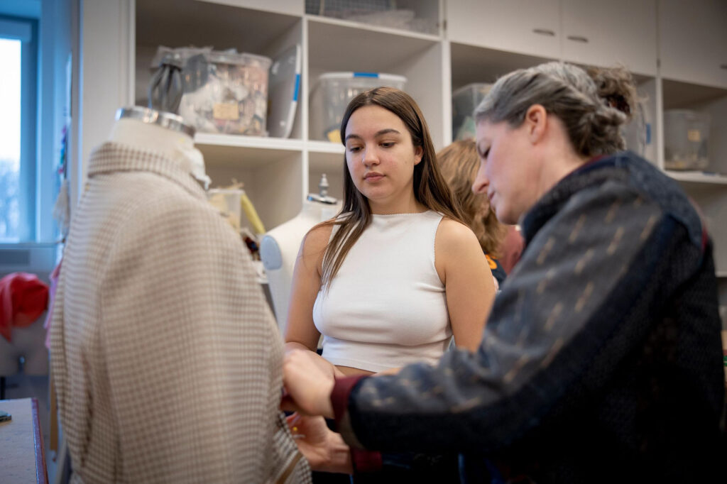 Two women sew a garment that is on a mannequin