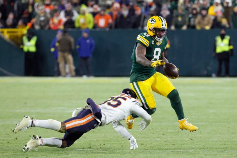 Green Bay Packers football players stand around a teammate lying on the field during a game, with stadium lights and spectators in the background.