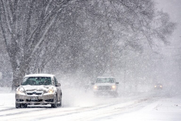 Three vehicles drive on a snow covered road during snowy conditions. The two cars in the distance can hardly be seen.