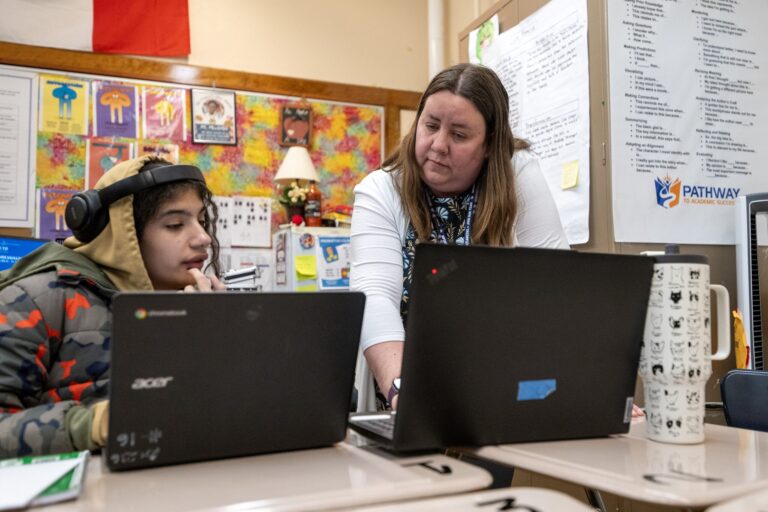 A teacher stands next to a student wearing headphones who is using a laptop in a classroom with other students working at desks.