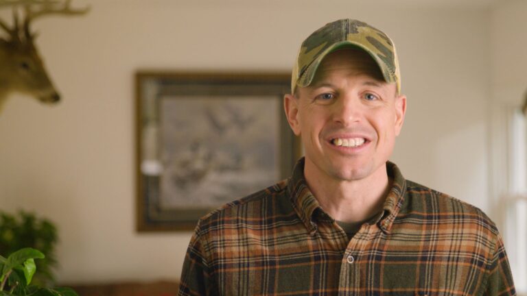 A man in a plaid shirt and camouflage cap smiles at the camera indoors, with a deer mount and framed picture in the background.