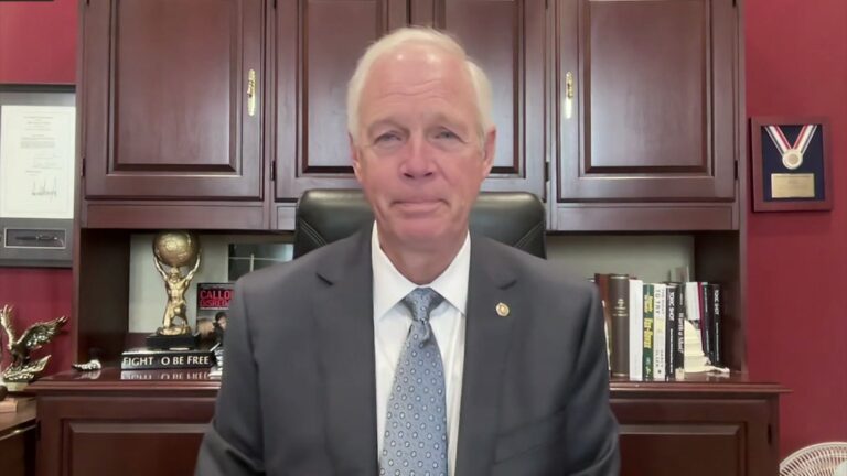 A man in a suit sits in an office with wooden cabinets, books, an eagle statue, and awards visible on the shelves behind him.