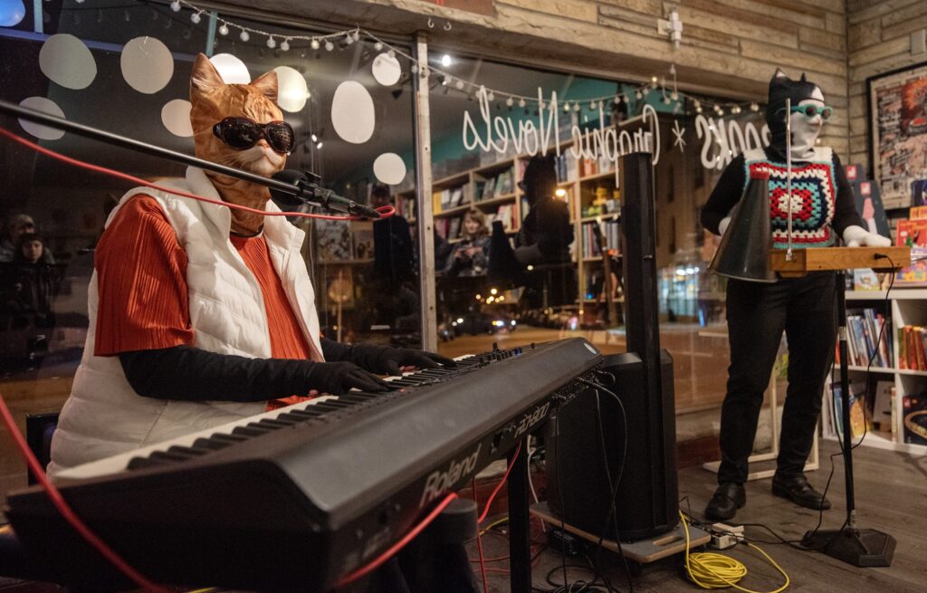 A person in a cat mask, crochet vest, and teal glasses uses a megaphone to address an audience in a room decorated with string lights and bookshelves.