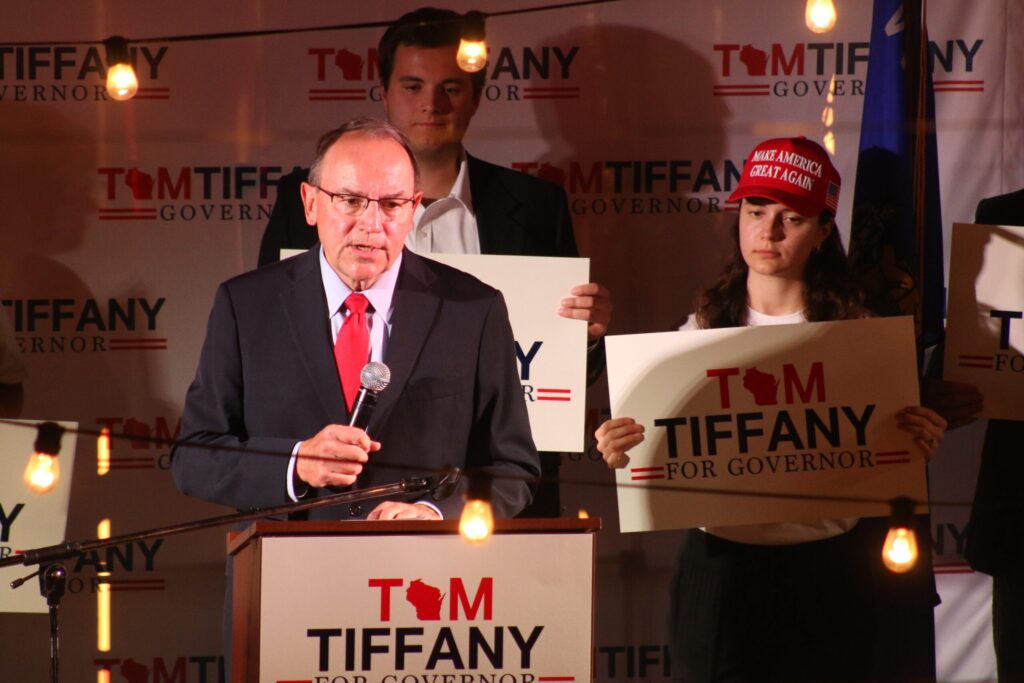 A man in a suit speaks at a podium with Tiffany for Governor signs behind him; supporters stand nearby, one wearing a Make America Great Again hat.
