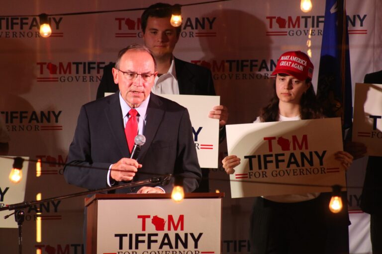 A man in a suit speaks at a podium with Tiffany for Governor signs behind him; supporters stand nearby, one wearing a Make America Great Again hat.