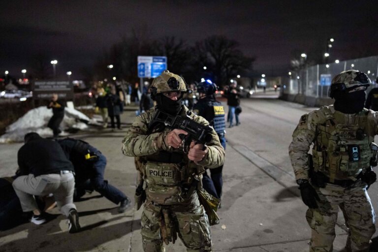 A woman is restrained against a car by several police and federal officers wearing tactical gear while bystanders observe and take photos.