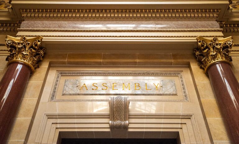 Ornate architectural detail above a doorway with the word ASSEMBLY engraved in gold letters, flanked by marble columns and decorative gold capitals.