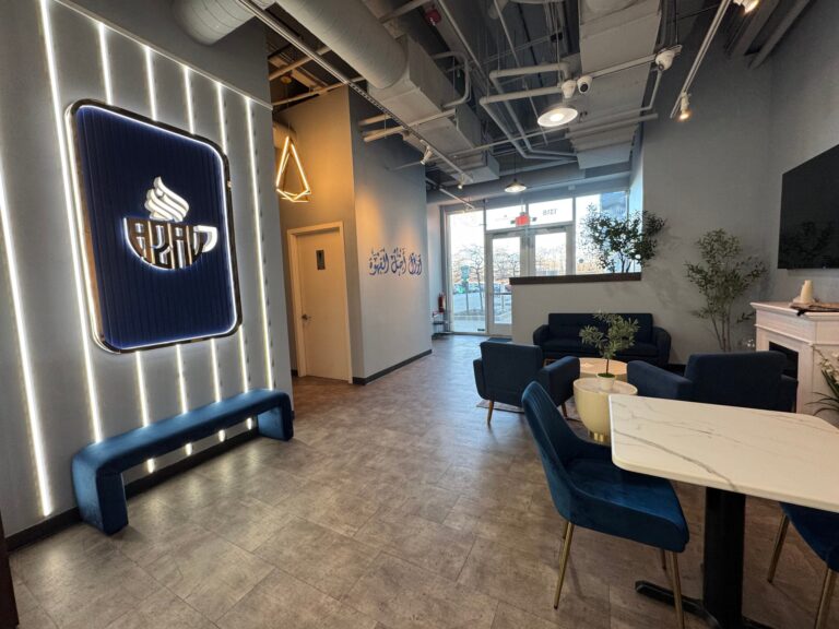 Modern cafe interior with blue seating, a marble table, wall signage, potted plants, and an illuminated cafe logo on the wall, with large windows letting in natural light.