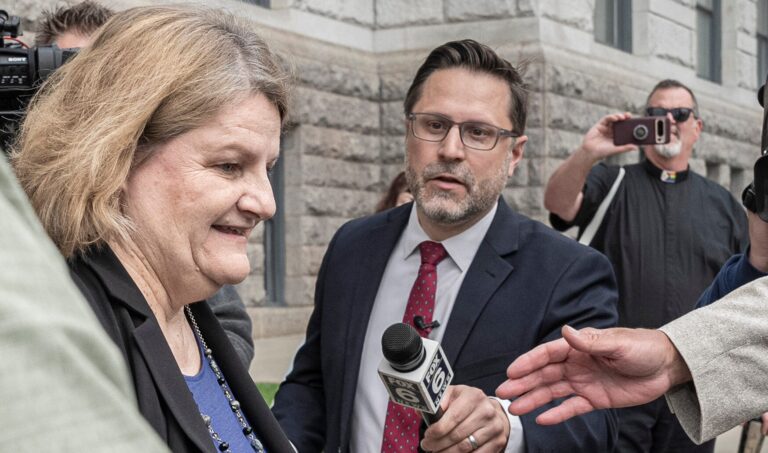 A group of people in winter clothing hold protest signs, including one that says We stand with Hannah, outside a building with arched stonework.