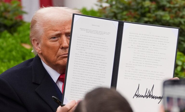 A person in a suit holds up a signed document in an outdoor setting, surrounded by greenery.