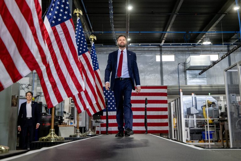 A man in a suit and red tie speaks at a podium with a Vice President of the United States seal in an industrial setting.