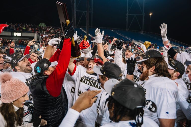 A football team celebrates on the field as a coach lifts a National Champions trophy, surrounded by cheering players in matching uniforms and hats.