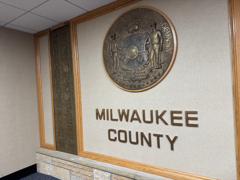 A man in a suit speaks at a podium in a formal government chamber with ornate lighting and arched windows. Other seated individuals are visible in the background.