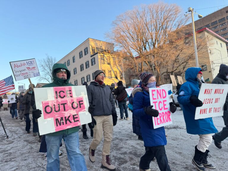 Five ICE officers wearing tactical gear and face coverings walk down a street beside parked vehicles on a cold day.
