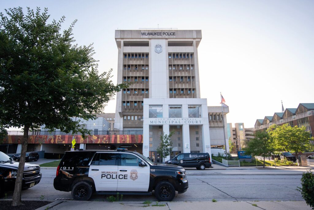 A Milwaukee Police SUV is parked in front of the Milwaukee Police Department headquarters and Municipal Court building on a sunny day.