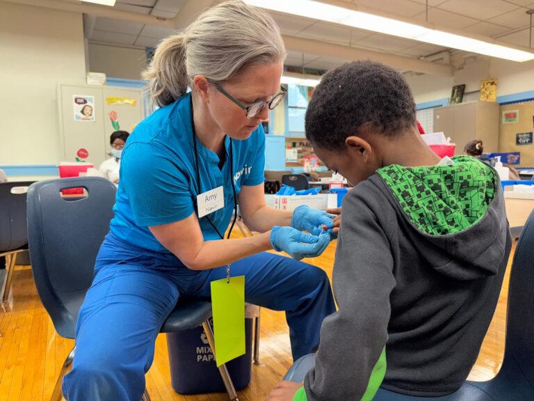 Containers with medical supplies, including syringes, alcohol pads, cotton swabs, and labeled vials, are organized on a wooden table in a clinical setting.