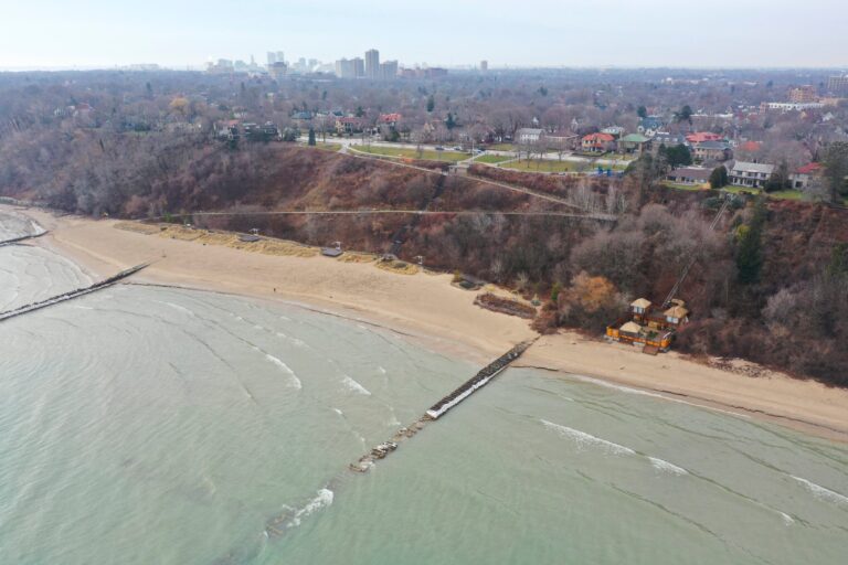 Aerial view of a sandy beach with wave breakers, adjacent to a tree-covered bluff and residential area, with a city skyline in the distance.