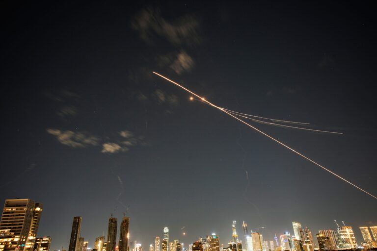 Long-exposure photo of rockets and interceptors crossing the night sky above a city skyline with illuminated buildings.