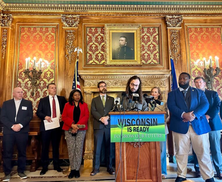 A group of people stand behind a podium with a Wisconsin Is Ready To Grow Our Future sign, speaking at a press event in an ornate room with a portrait on the wall.