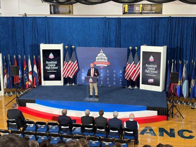 Two men pose in front of an Education 250 branded backdrop with U.S. flags at a formal event.