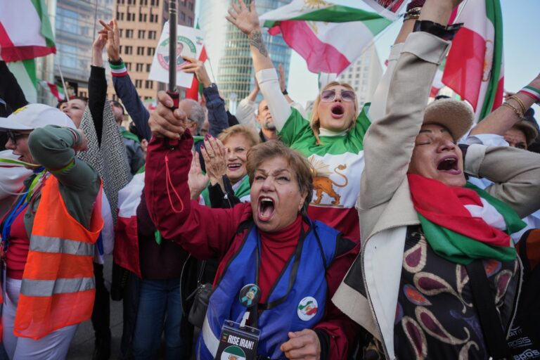 A group of people, some wearing flags and vests, stand close together, raising their arms and shouting or cheering during a public gathering or protest.