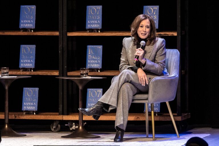 A stage event features Kamala Harris in conversation with a moderator, with a large screen displaying 107 Days and event details in the background.