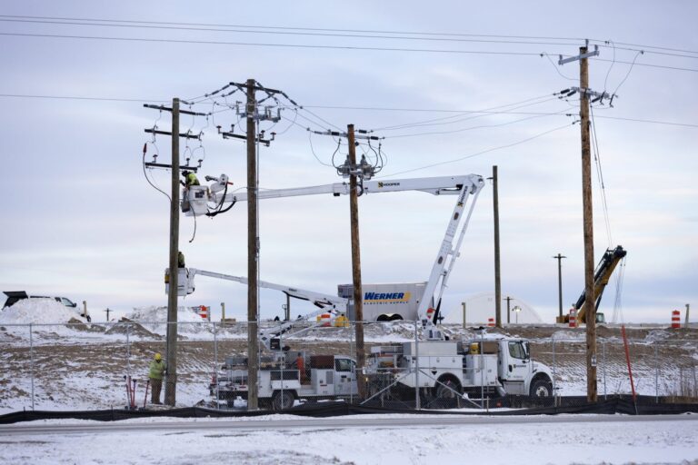 High-voltage power lines and utility poles stretch across a landscape at dusk, with a gradient sky and silhouetted trees in the background.