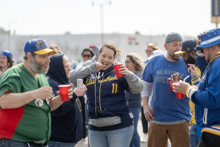 A man grills burgers and sausages on a red barbecue in a parking lot while others stand nearby, tailgating before an event.