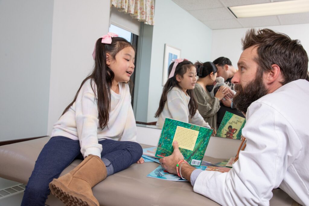 A child’s hands are spread across a pile of colorful children’s books on a table, some partially overlapping each other.