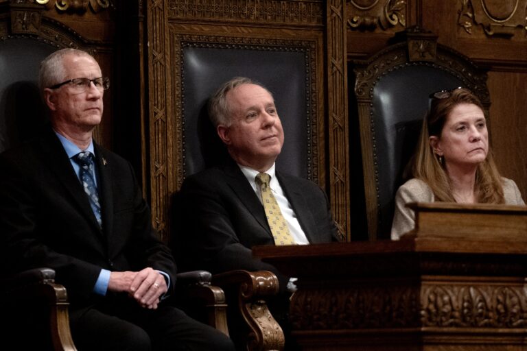 Three people in formal attire sit side by side in ornate chairs in a wood-paneled room, appearing attentive and serious.