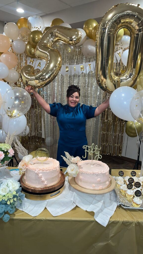 A person in winter clothing stands under a wooden arch decorated with white and blue string lights in an outdoor nighttime setting.