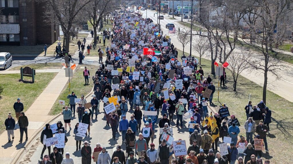 Large crowd of people march down a city street holding signs and banners during a protest on a clear day.