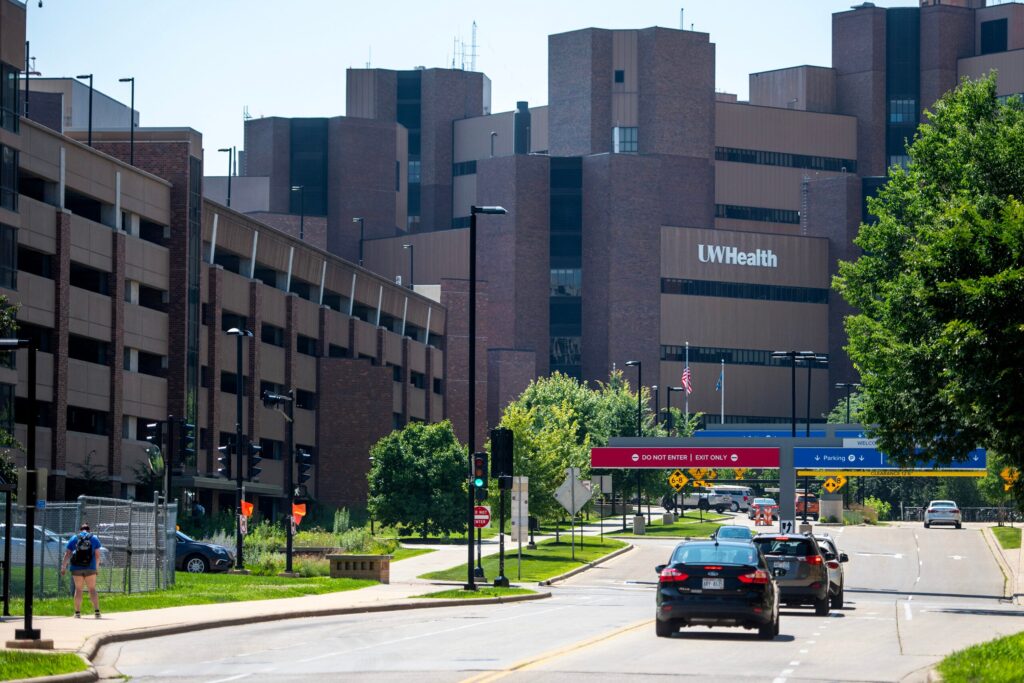 Several cars drive towards the UW Health building, with a parking structure and pedestrian visible along the street on a sunny day.