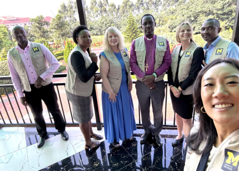 Seven people standing on a balcony take a selfie with trees and foliage behind them