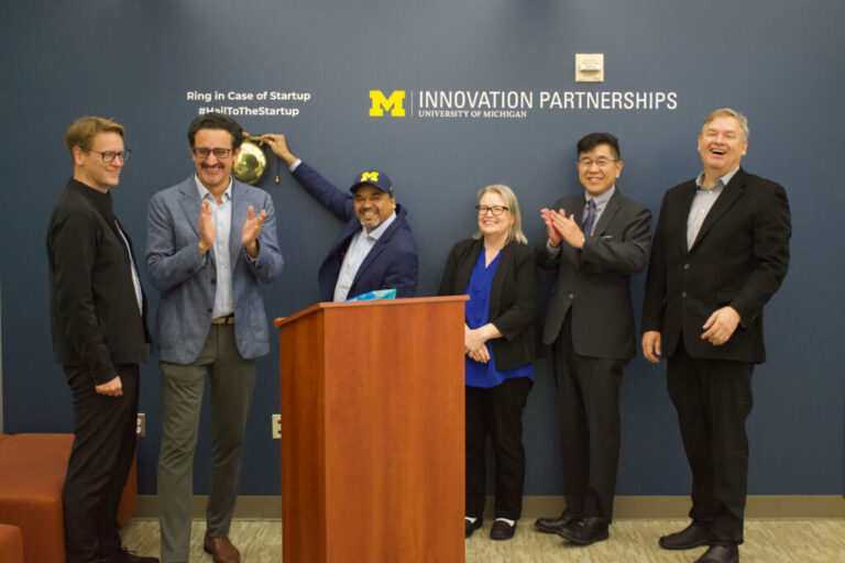Six people standing behind a podium, ringing a bell on the wall, in celebration