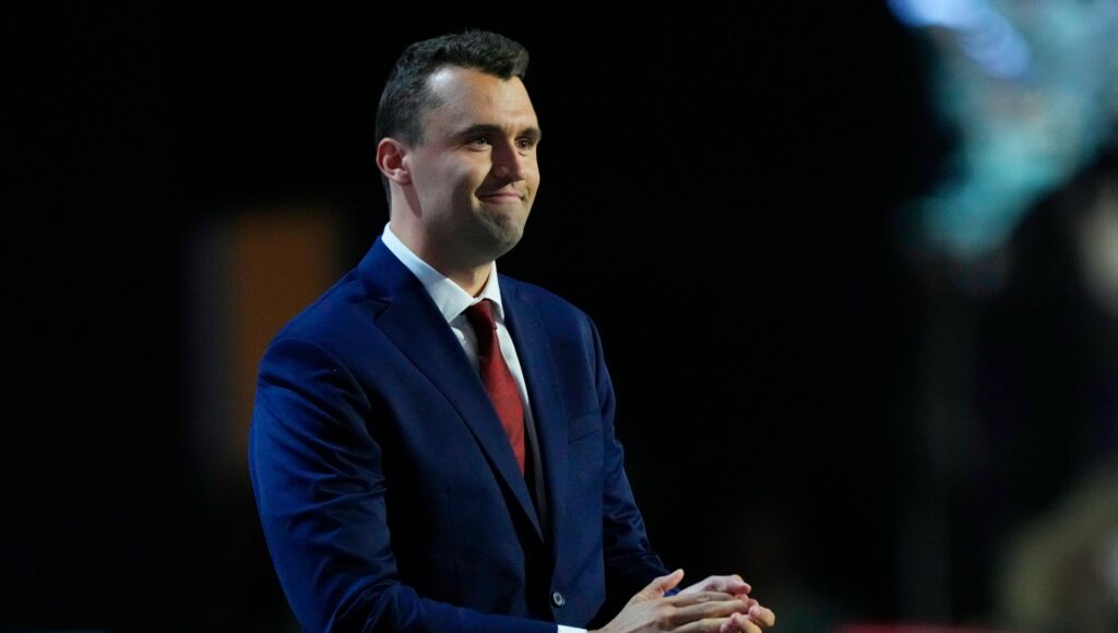 A man in a dark blue suit and red tie stands on stage, clasping his hands, with a dark background and blurred audience.