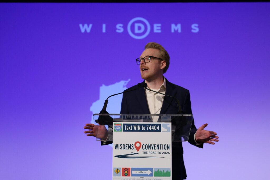 A person speaks at a podium during the WisDems Convention, with a microphone and a projected WisDems logo in the background.