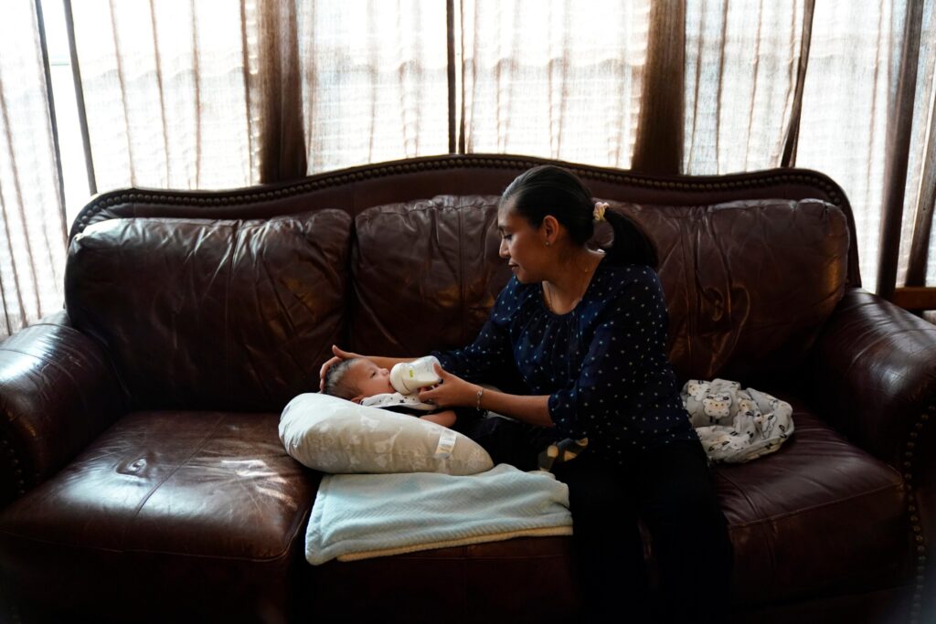 A woman sits on a brown leather couch, bottle-feeding a baby who is lying on a pillow with a blanket.
