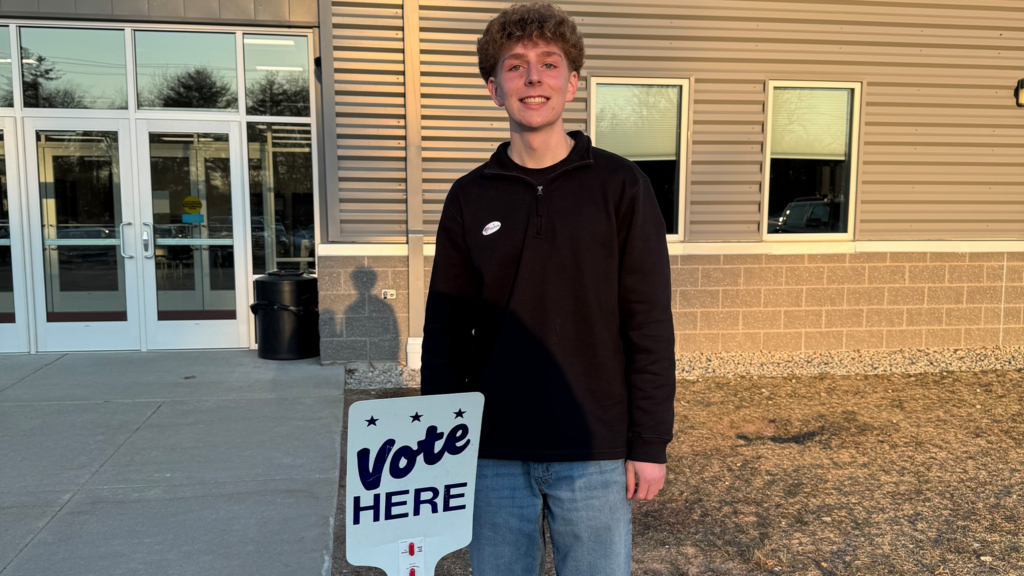 A young person stands outside a building next to a Vote Here sign, wearing a sticker, with sunlight casting shadows on the ground.